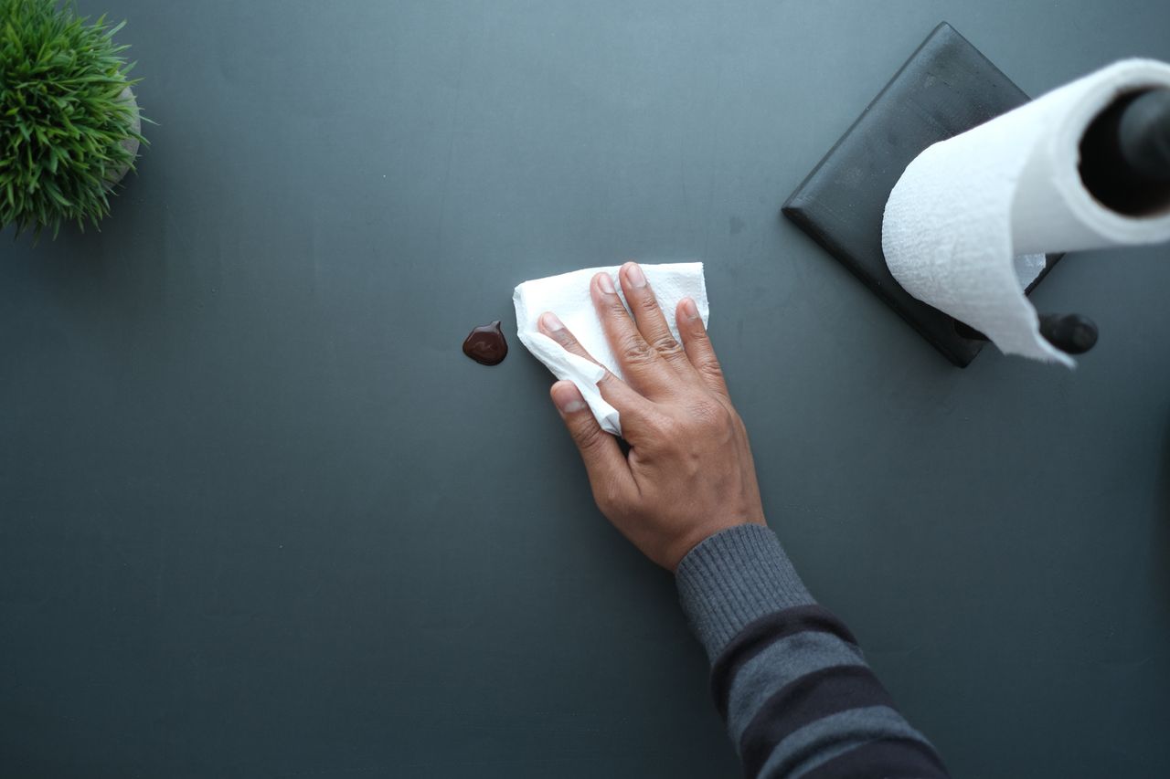 heros-img A hand wipes a dark surface with a tissue near a paper roll and plant.
