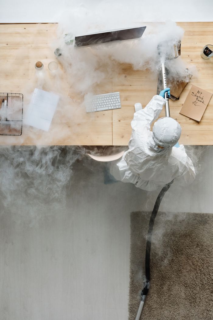 about-us-02 A person in protective gear sanitizing an office desk using fumigation equipment during daytime.